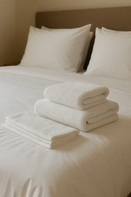 Opgemaakt bed met witte lakens en een keurig gestapelde set hotelhanddoeken op de voorgrond.