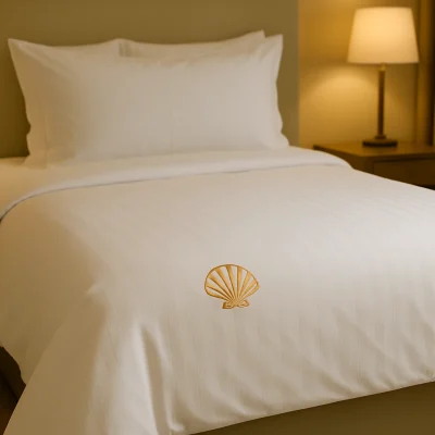 Wit hotel dekbedovertrek met een goudkleurig schelp-logo geborduurd aan het voeteneind, opgemaakt op een luxe hotelbed.