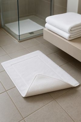 Witte hotelbadmat op grijze tegels; hoek omgeslagen met zichtbare antislip backing.