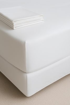 Close-up van bedhoek met strak wit hoeslaken; matte percale-textuur en gevouwen lakens bovenop.